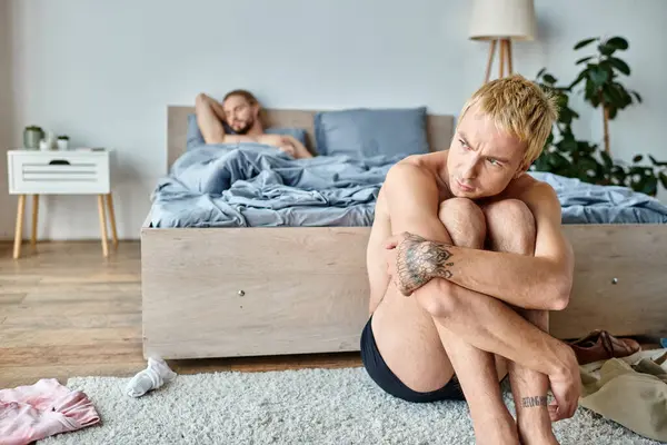 Offended tattooed gay man sitting near love partner lying in bedroom in morning, troubled love — Stock Photo