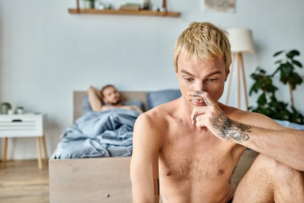 Upset tattooed gay man touching face near love partner lying in bedroom in morning, troubled love — Stock Photo