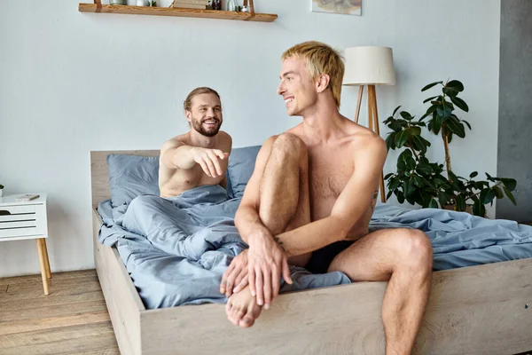 Счастливый бородатый гей с протянутой рукой рядом с улыбающимся парнем, сидящим на кровати утром — стоковое фото