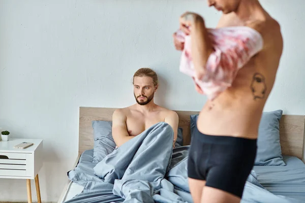 Upset bearded gay man sitting on bed near love partner dressing up in bedroom, relationship issues — Stock Photo