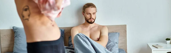 Засмучений бородатий гей чоловік сидить на ліжку поруч з любовним партнером, одягненим у спальню, горизонтальний банер — стокове фото