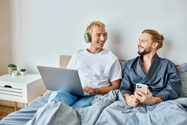 Happy gay man in headphones browsing internet on laptop near boyfriend with smartphone on bed — Stock Photo