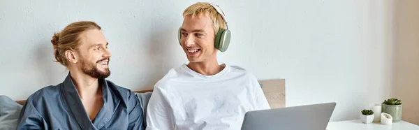 Happy gay man in headphones smiling at bearded boyfriend near laptop in bedroom, horizontal banner — Stock Photo