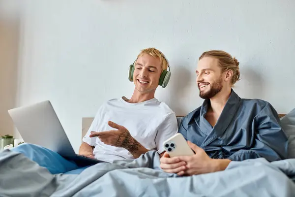 Happy gay man in headphones pointing at laptop near bearded boyfriend with smartphone in bedroom — Stock Photo