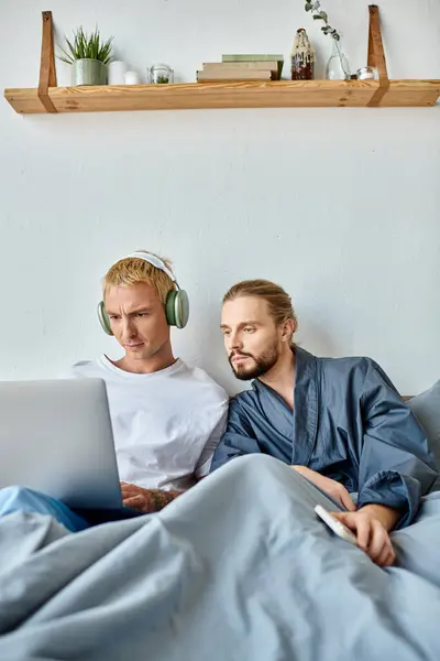 Serious gay man in headphones networking on laptop near bearded boyfriend with smartphone in bedroom — Stock Photo