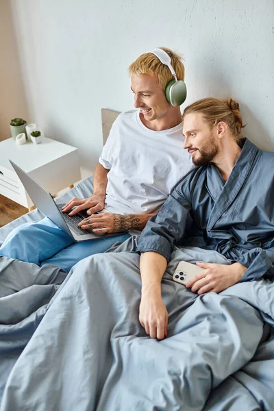 Cheerful gay man in headphones using laptop near bearded boyfriend with smartphone in bedroom — Stock Photo