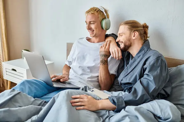 Alegre gay homem no fones de ouvido usando laptop perto barbudo namorado com smartphone no quarto — Fotografia de Stock
