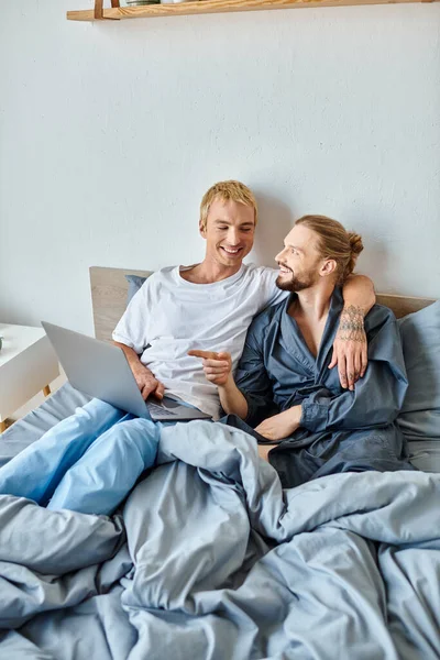 Веселый бородатый гей указывает на ноутбук во время просмотра фильма на ноутбуке с парнем на кровати — стоковое фото