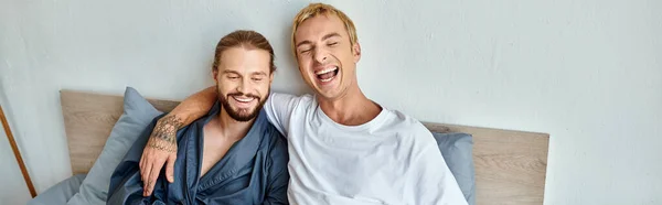 Cheerful gay couple laughing while sitting on bed in morning, happy relationships, horizontal banner — Stock Photo