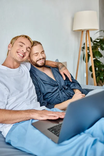 Delighted gay couple sitting with closed eyes while watching romantic movie on laptop in bedroom — Stock Photo