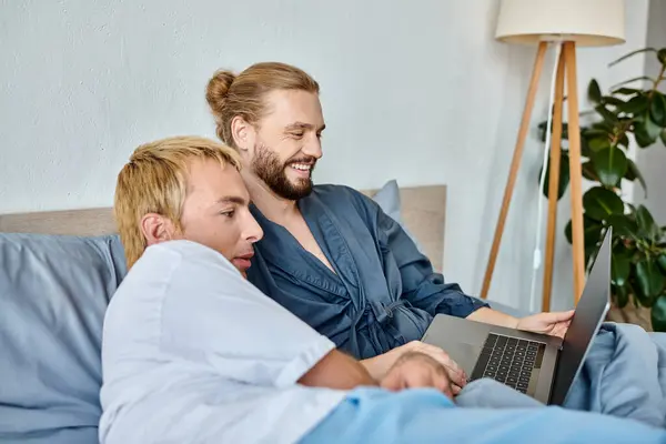 Радісний бородатий гей-чоловік посміхається під час перегляду комедійного фільму на ноутбуці з хлопцем у спальні — стокове фото