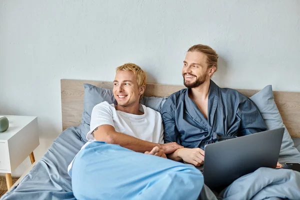 Щаслива гей пара дивиться далеко під час перегляду фільму на ноутбуці в спальні, дозвіллі та спокої — стокове фото