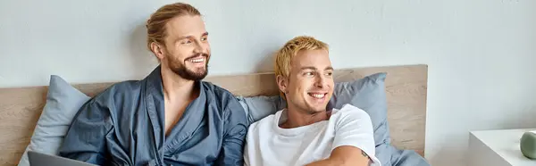 Happy gay couple smiling and looking away while sitting in bedroom in morning, horizontal banner — Stock Photo