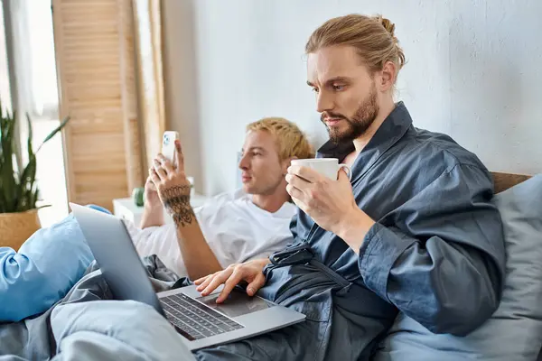 Barbu gay homme avec café tasse réseau sur ordinateur portable près tatoué copain avec smartphone sur lit — Photo de stock