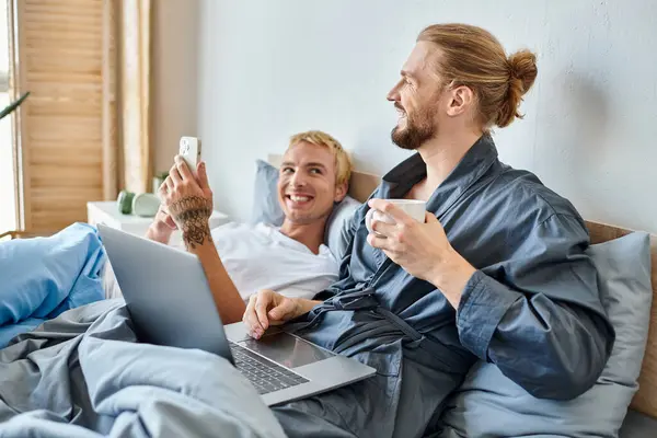 Barbuto gay uomo con caffè tazza sorridente vicino laptop e tatuato fidanzato con smartphone su letto — Foto stock