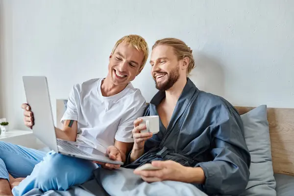 Веселий гей-чоловік показує ноутбук усміхненому бородатому хлопцеві, який п'є ранкову каву в спальні — стокове фото