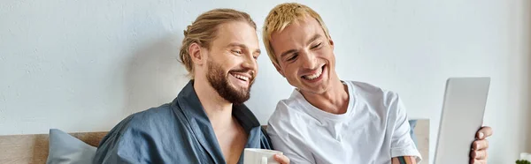 Cheerful gay man showing laptop to smiling bearded boyfriend drinking coffee in bedroom, banner — Stock Photo