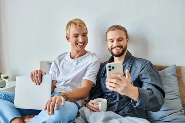 Joyful gay man with coffee cup showing smartphone to smiling boyfriend with laptop in bedroom — Stock Photo
