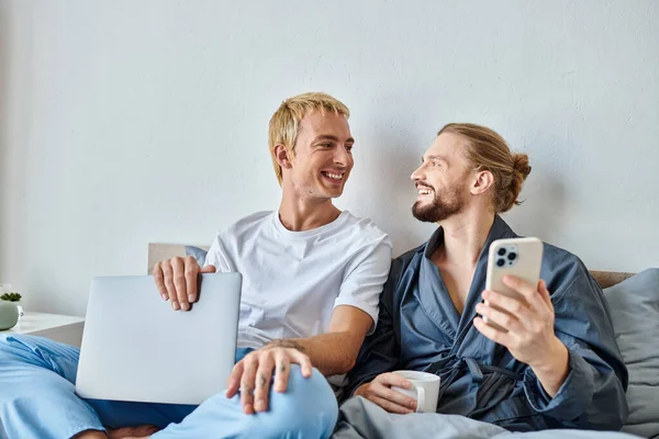 Joyful gay man with coffee cup and smartphone smiling at boyfriend with laptop in bedroom, leisure — Stock Photo