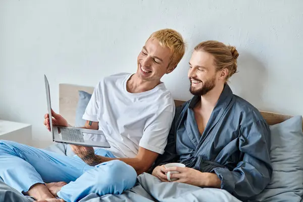 Joyful gay man showing laptop to smiling bearded man sitting with coffee cup in bedroom, leisure — Stock Photo