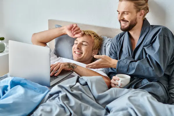Happy tattooed gay man browsing internet on laptop near bearded boyfriend with coffee cup in bedroom — Stock Photo