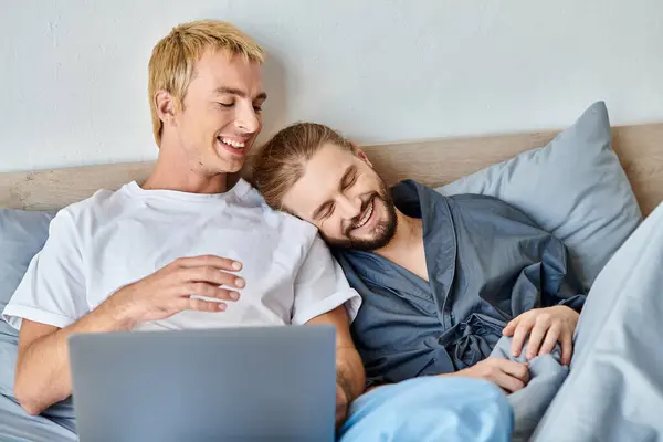 Улыбающаяся гей-пара смотрит романтический фильм на ноутбуке в спальне, счастливого утреннего отдыха на кровати — стоковое фото