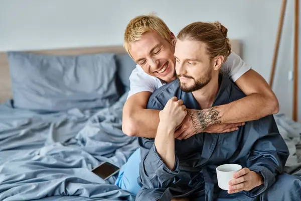 Freudig tätowiert Homosexuell Mann umarmt bärtigen Freund mit Kaffeetasse im Schlafzimmer, glückliche Szene — Stockfoto