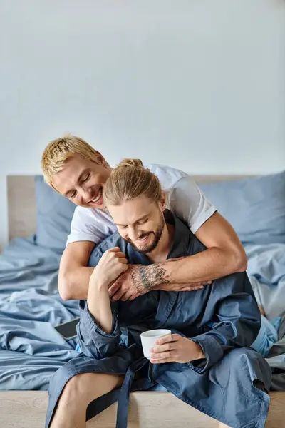 Joyeux tatoué gay homme embrassant barbu copain avec tasse de café dans chambre, scène heureuse — Photo de stock