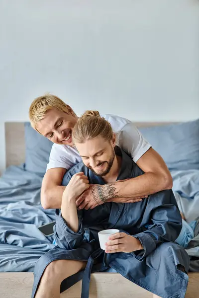Joyful tattooed gay man embracing bearded boyfriend with coffee cup in bedroom, happy scene — Stock Photo