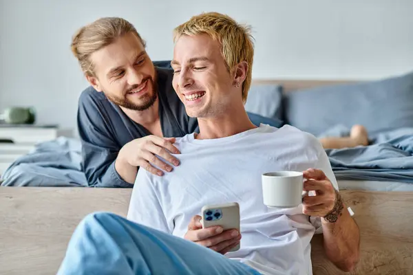 Веселый бородатый гей улыбается рядом с парнем с чашкой кофе с помощью мобильного телефона в спальне — стоковое фото