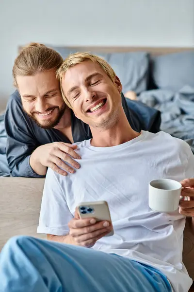 Весёлый бородатый гей улыбается рядом с парнем с чашкой кофе и мобильным телефоном в спальне — стоковое фото