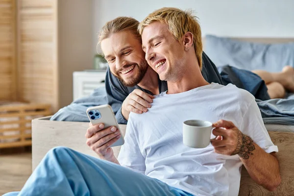 Веселый бородатый гей улыбается рядом с парнем с чашкой кофе с помощью мобильного телефона в спальне — стоковое фото