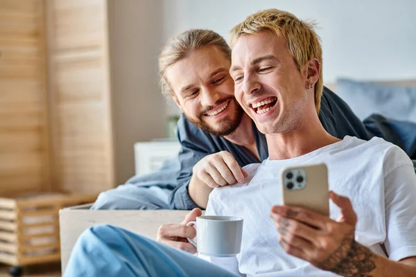 Cheerful tattooed gay man with coffee cup browsing internet on smartphone near smiling boyfriend — Stock Photo