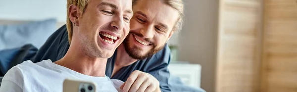 Alegre tatuado gay homem navegação internet no smartphone perto sorrindo namorado no quarto, banner — Fotografia de Stock