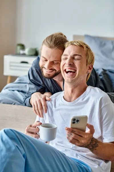 Laughing gay man with coffee cup browsing internet on mobile phone near smiling boyfriend in bedroom — Stock Photo