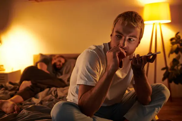 Desleal gay homem envio voz mensagem no smartphone perto namorado dormindo no noite no quarto — Fotografia de Stock
