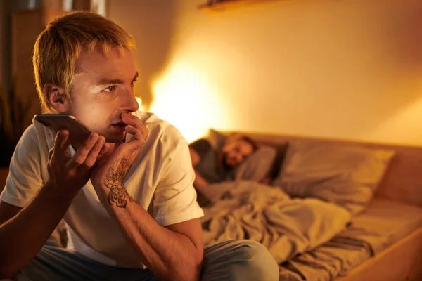Tattooed disloyal gay man sending voice message on smartphone near boyfriend sleeping in bedroom — Stock Photo