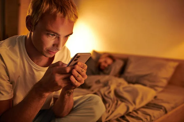 Disloyal gay man messaging on mobile phone near boyfriend sleeping at night in bedroom, cheating — Stock Photo