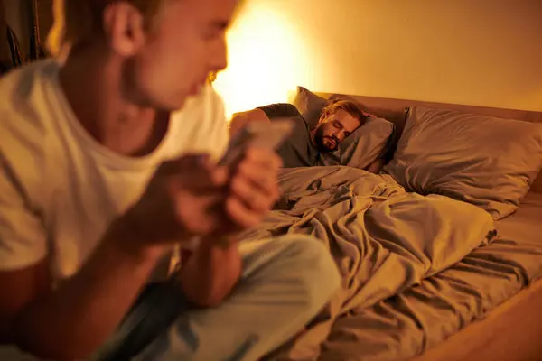 Gay man sleeping at night while unfaithful boyfriend chatting on smartphone on blurred foreground — Stock Photo