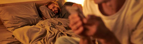 Gay hombre durmiendo en la noche cerca infiel novio con smartphone en borrosa primer plano, bandera - foto de stock