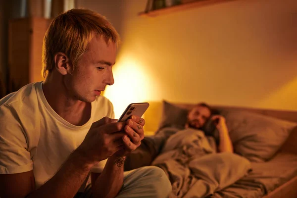 Disloyal gay man messaging on mobile phone near boyfriend sleeping at night in bedroom, cheating — Stock Photo