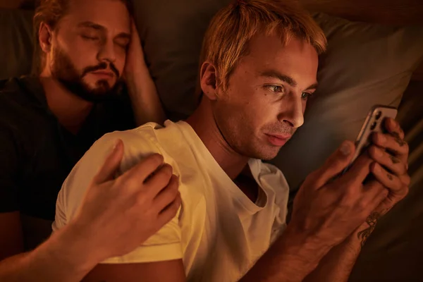 Disloyal gay man browsing date app on mobile phone near boyfriend sleeping at night in bedroom — Stock Photo