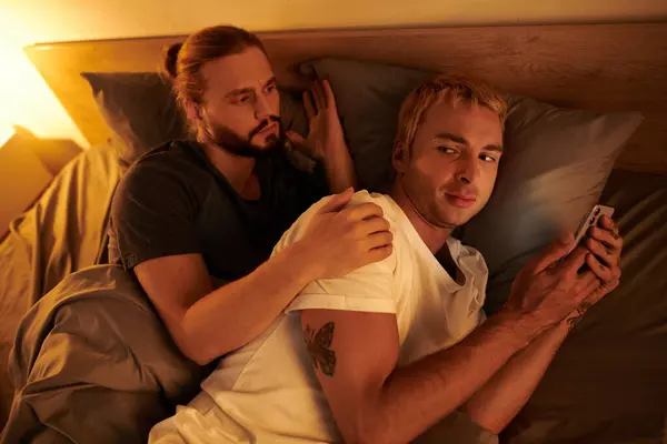 Disloyal gay man browsing date app on mobile phone near discouraged boyfriend at night in bedroom — Stock Photo