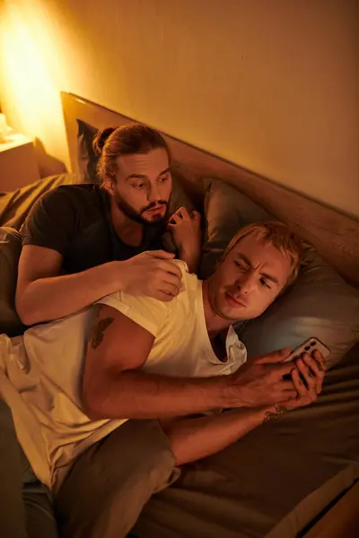 Unfaithful gay man browsing date app on mobile phone near discouraged boyfriend at night in bedroom — Stock Photo