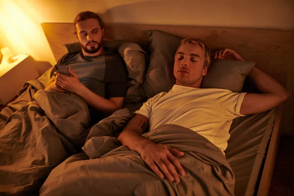 Infiel barbudo gay mensajería en móvil cerca durmiendo novio en la noche en dormitorio - foto de stock