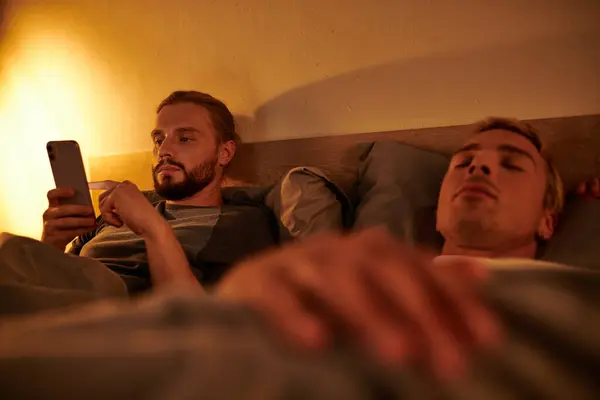 Unfaithful bearded gay messaging on mobile phone near sleeping boyfriend at night in bedroom — Stock Photo