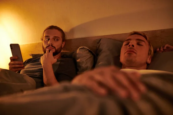 Unfaithful bearded gay browsing date app on smartphone near sleeping boyfriend at night in bedroom — Stock Photo