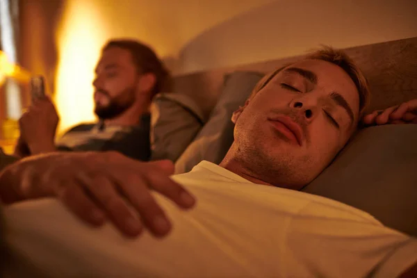 Gay man sleeping near unfaithful boyfriend browsing date app on smartphone at night in bedroom — Stock Photo