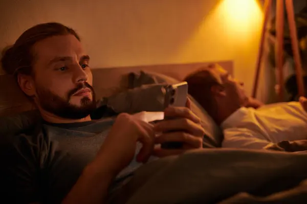 Unfaithful bearded gay man messaging on mobile phone near sleeping boyfriend at night in bedroom — Stock Photo