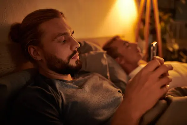 Unfaithful bearded gay man messaging on mobile phone near sleeping boyfriend at night in bedroom — Stock Photo